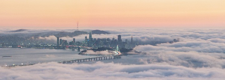 A cityscape with a bridge is partially obscured by a thick layer of fog under a pastel sky.