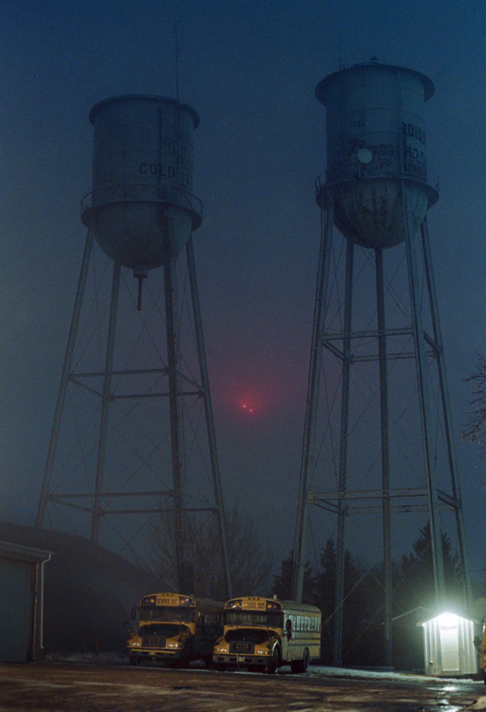 Two school buses are parked in front of two water towers on a foggy night.