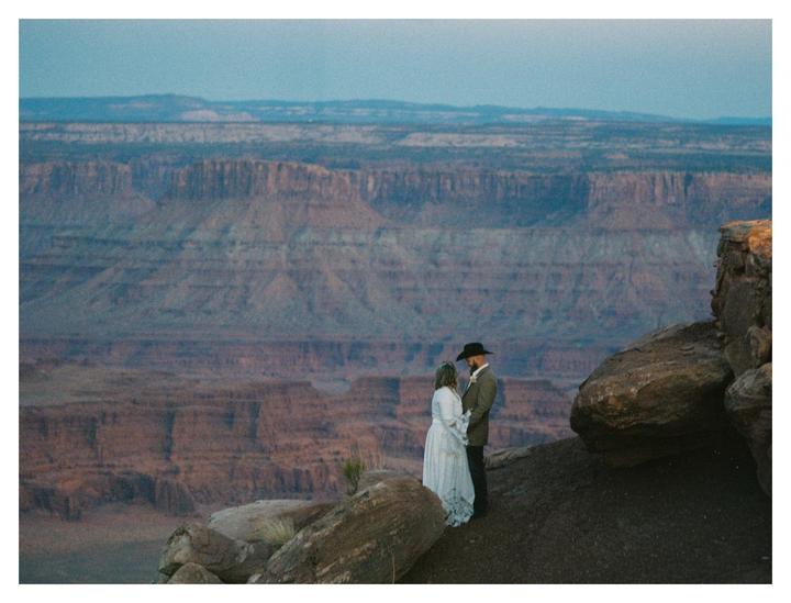 A couple stands on a cliff overlooking a vast canyon landscape.