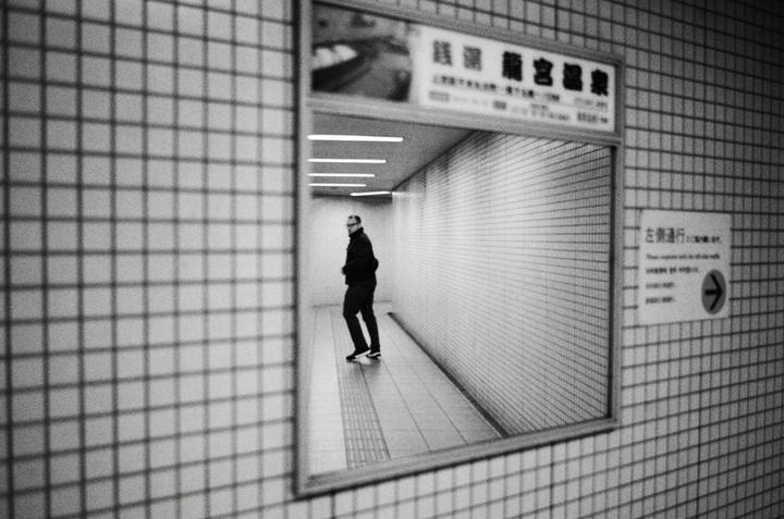 A man walks down a tiled hallway, reflected in a mirror on a tiled wall.