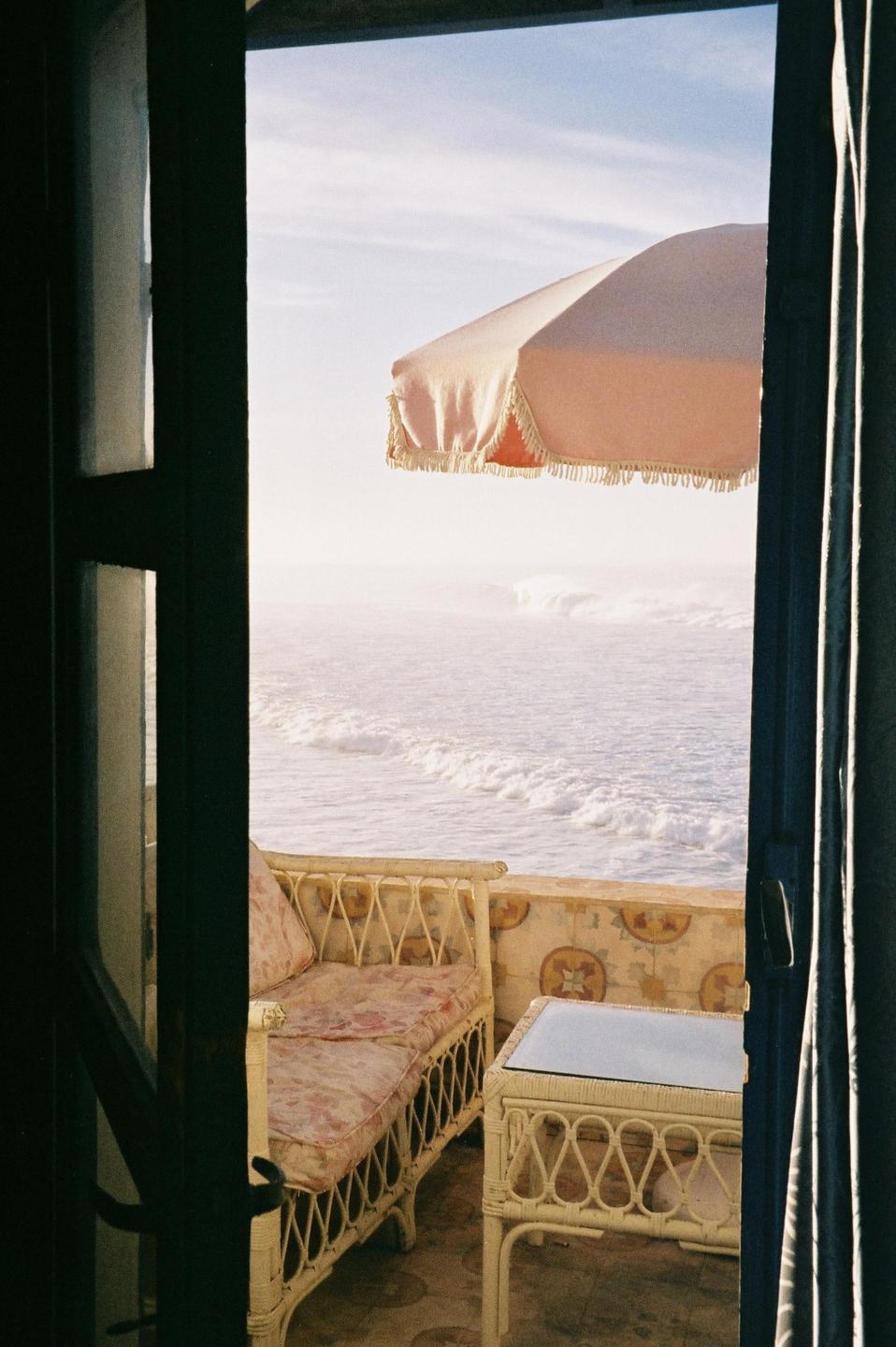A balcony with a wicker sofa and table overlooks a hazy ocean with waves and a pink umbrella.