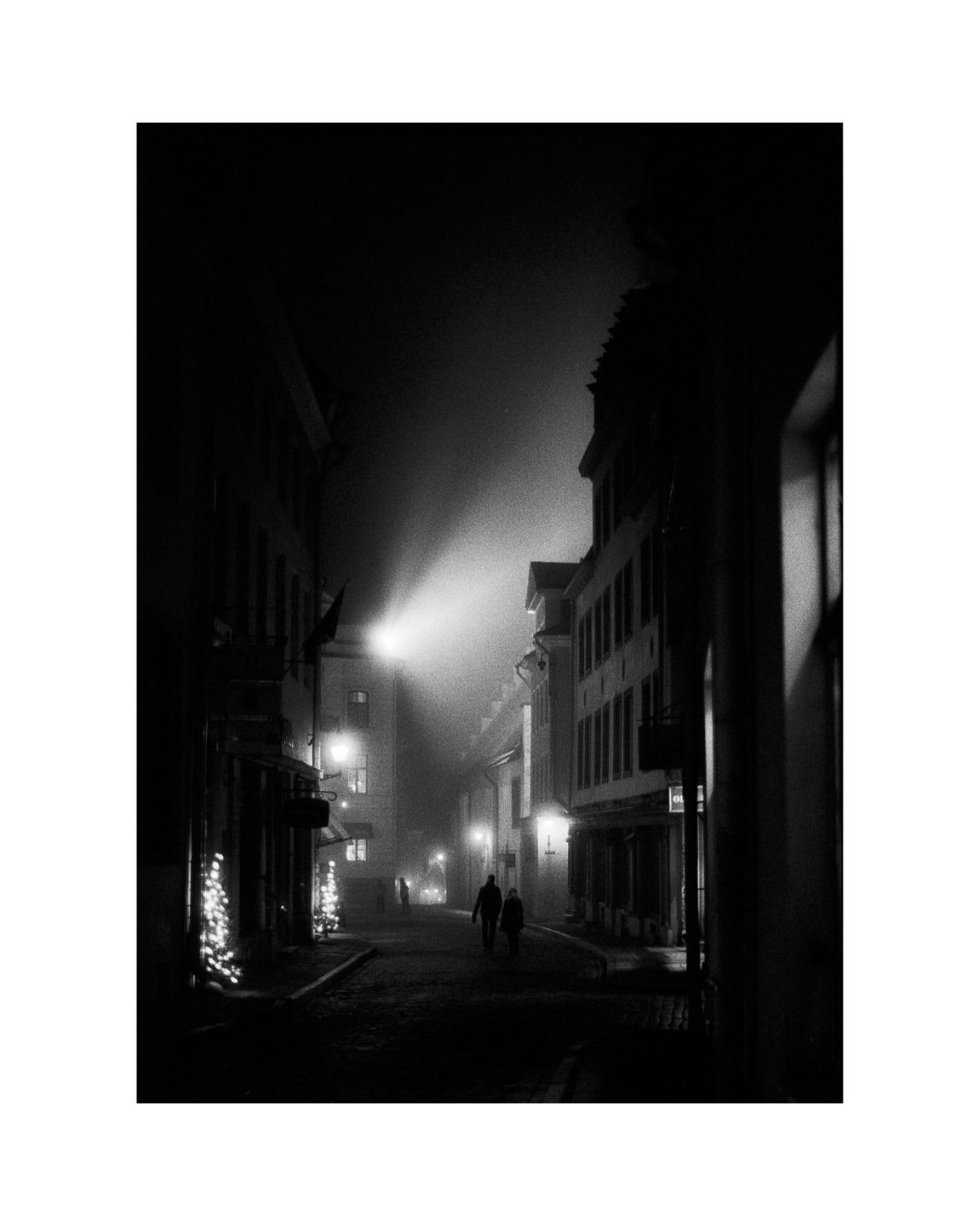 A couple walks down a foggy street lined with buildings and lights at night.