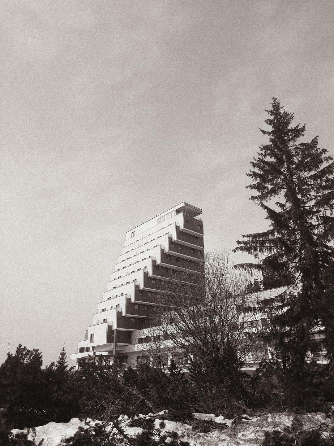 A tall, stepped building stands among trees and snow under a bright sky.