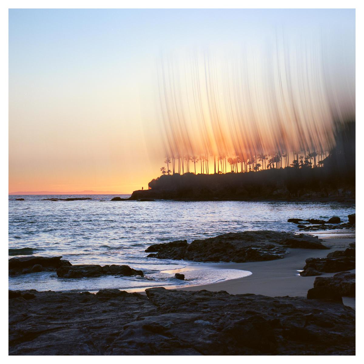 The sun sets behind a line of palm trees on a rocky beach, creating a streaky light effect.