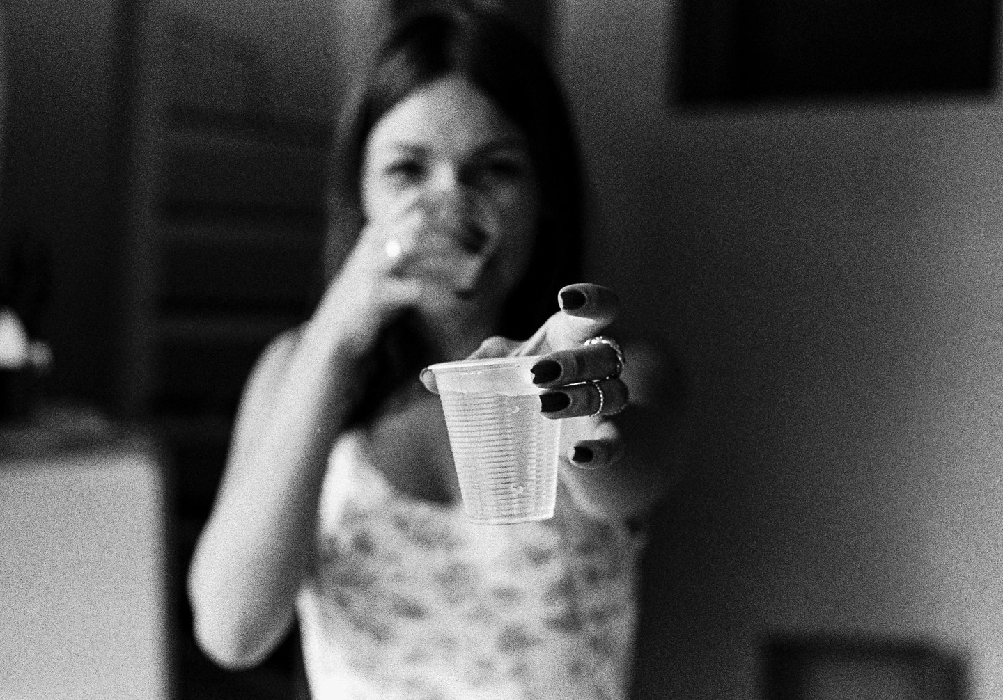 A woman extends a plastic cup toward the viewer while holding another cup to her mouth.
