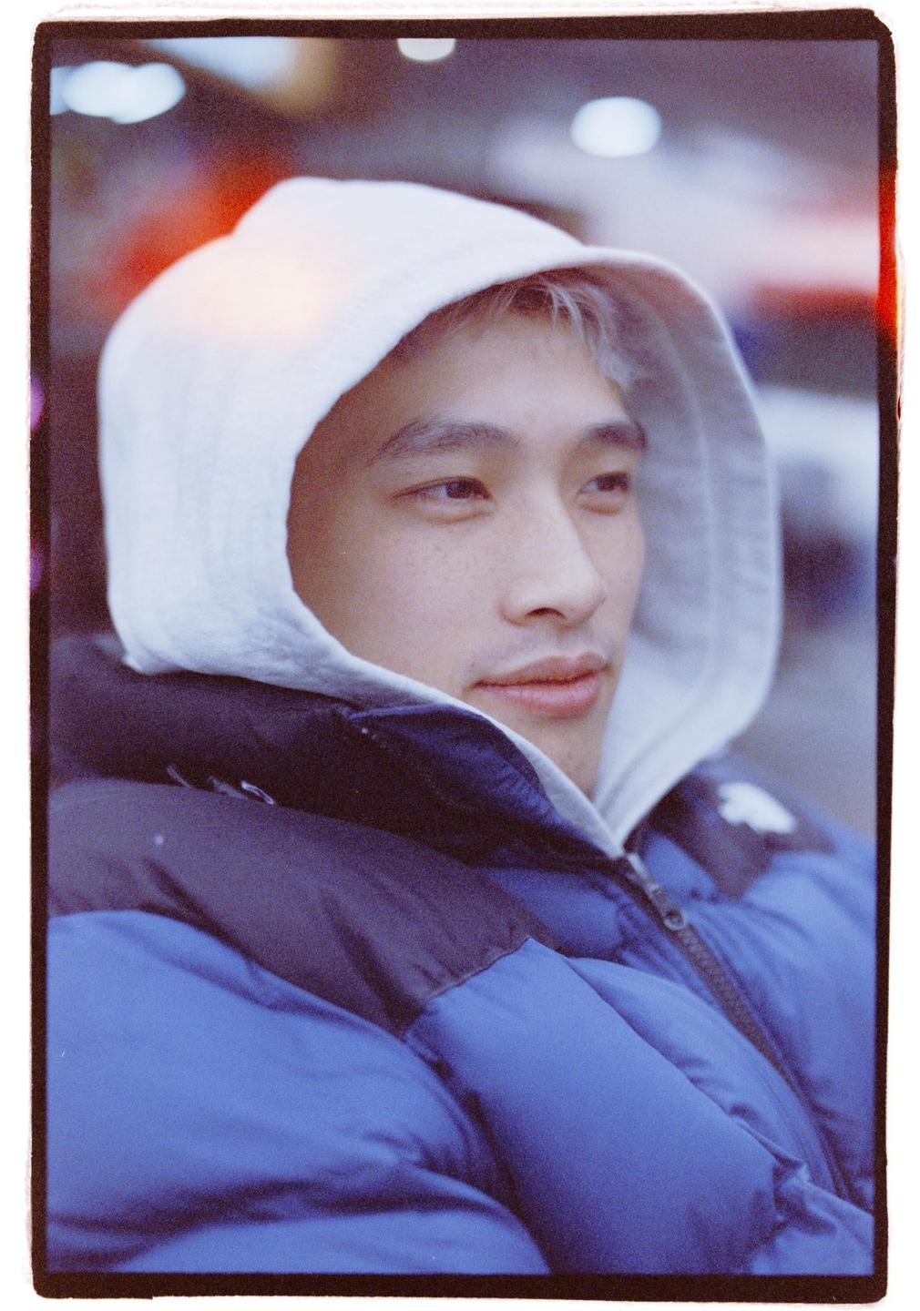 A man in a blue puffer jacket and a white hoodie looks to the right.