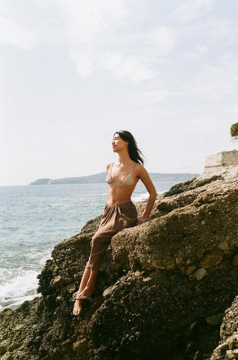 A woman in a bikini top and pants sits on a rocky shore looking out at the ocean.