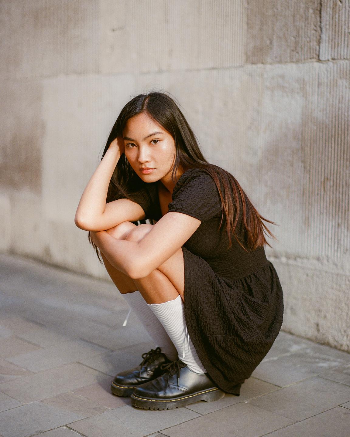 A woman in a black dress and white socks crouches on a paved surface next to a stone wall.