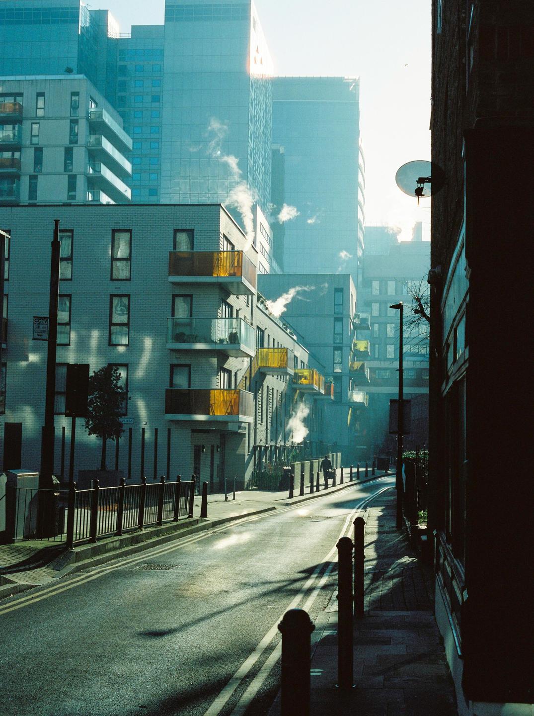 A street in East London is lined with modern buildings, some with yellow balconies, and steam rises from several places.