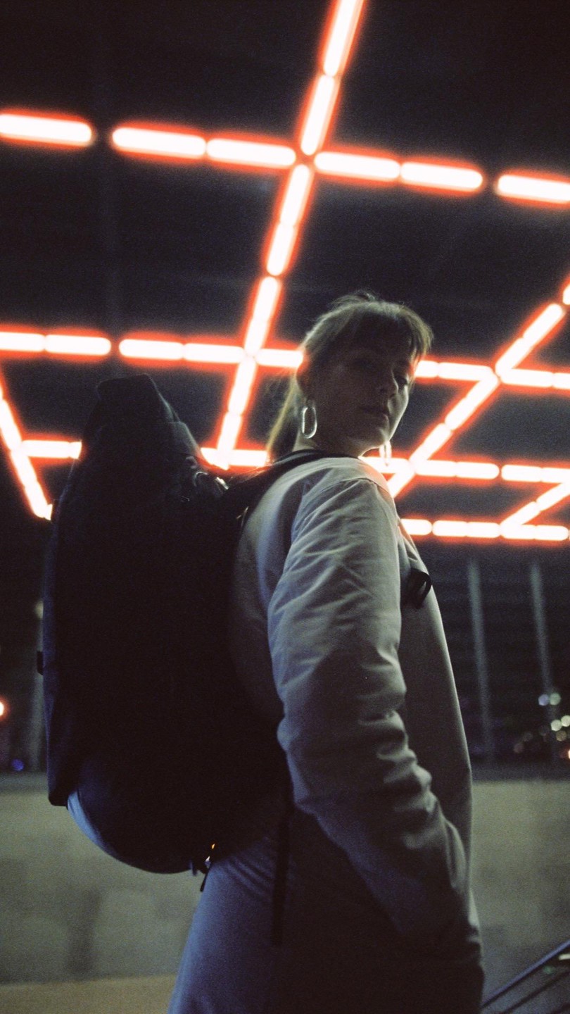 A woman with a backpack stands in front of a neon light installation at night.