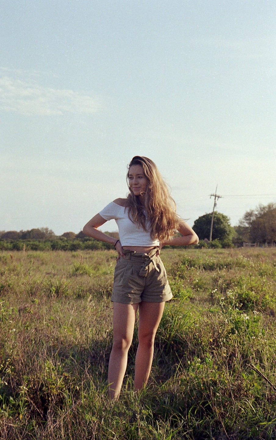 A woman with long hair stands in a grassy field with her hands on her hips, looking to the side.