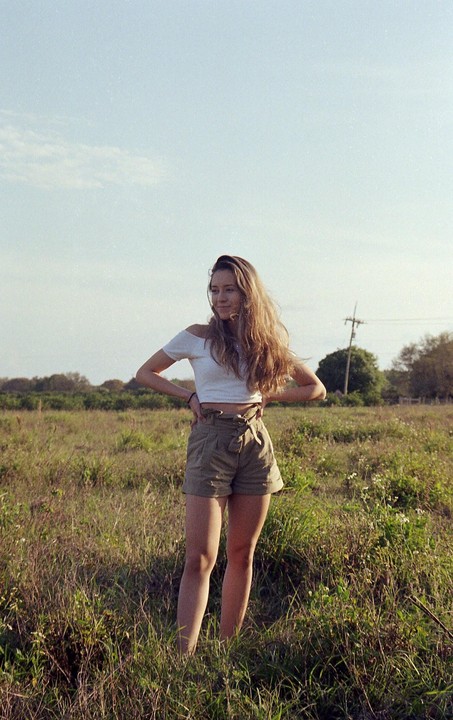 A woman with long hair stands in a grassy field with her hands on her hips, looking to the side.
