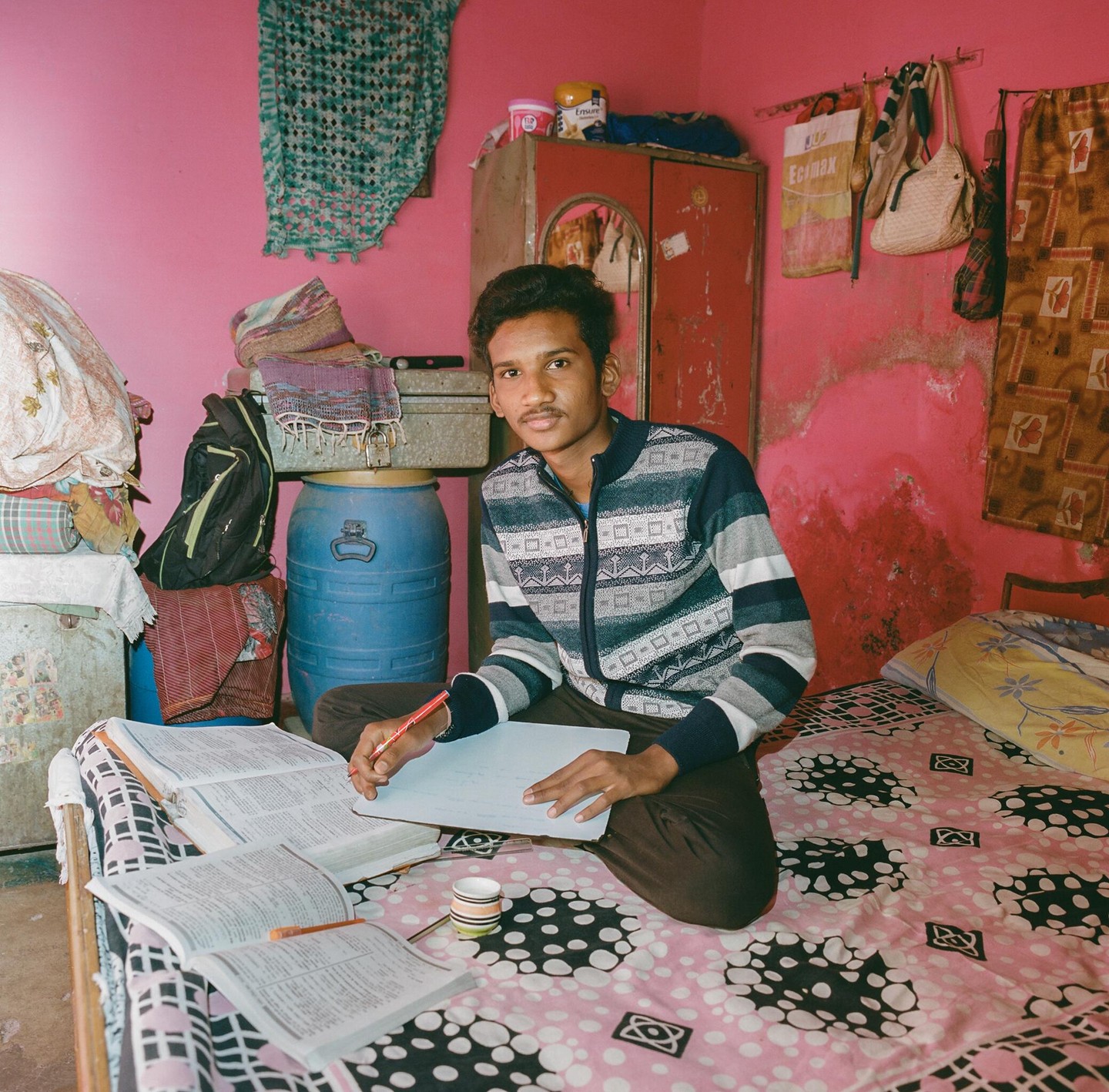 A young man sits on a bed with open books and a notebook, holding a pencil, in a room with pink walls.
