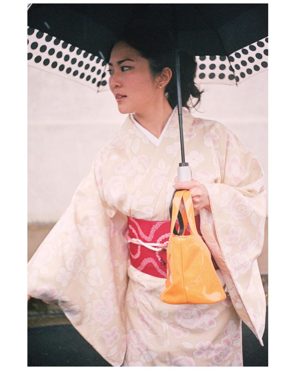 A woman in a kimono holds a polka dot umbrella and an orange handbag.