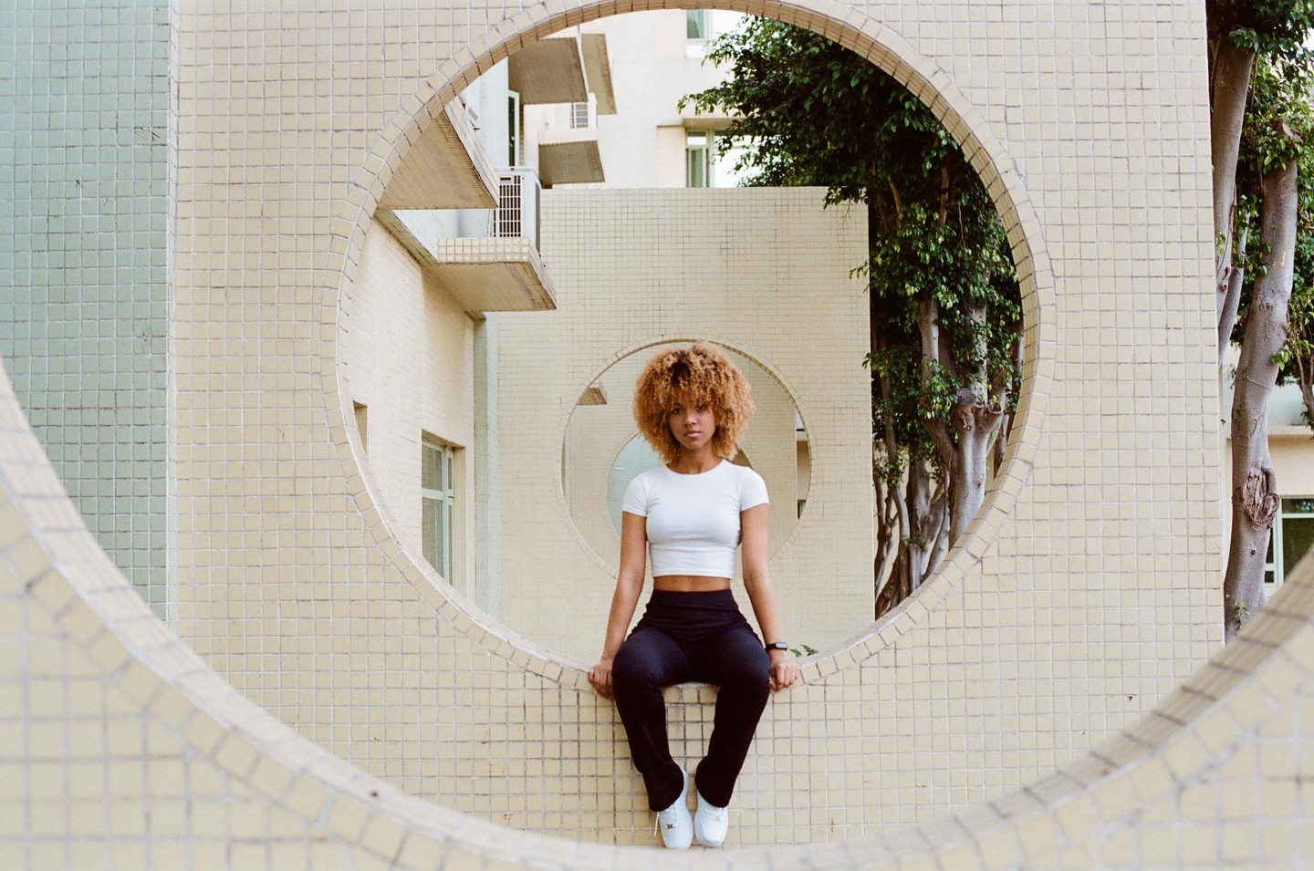 A woman with curly hair sits in a circular opening of a tiled wall.