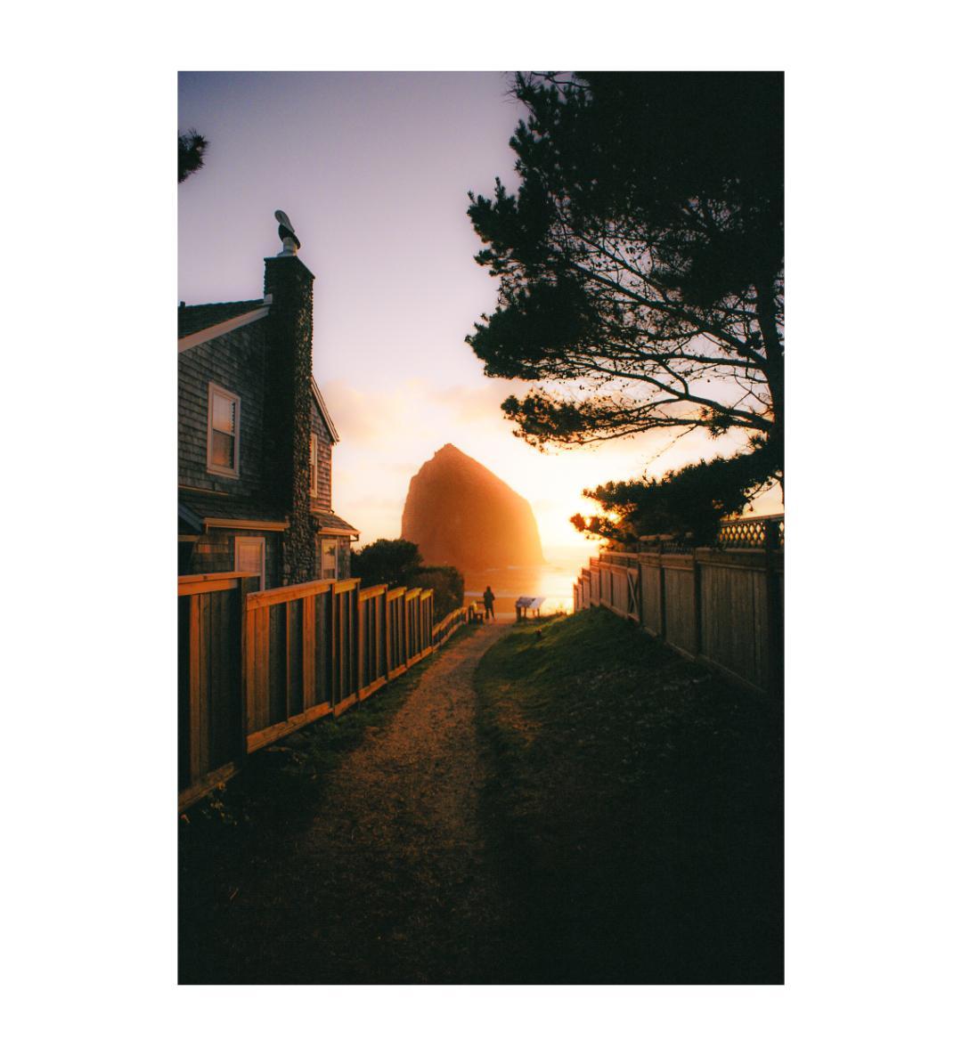 A path leads to a large rock formation on a beach at sunset, with a house and fence on the left and a tree on the right.