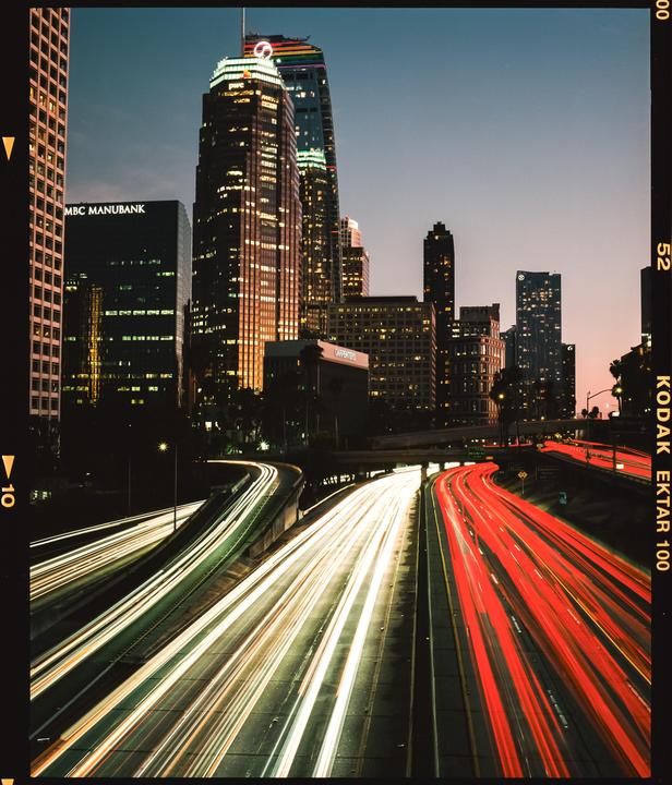 Light trails from vehicles on a highway lead towards a city skyline at dusk.