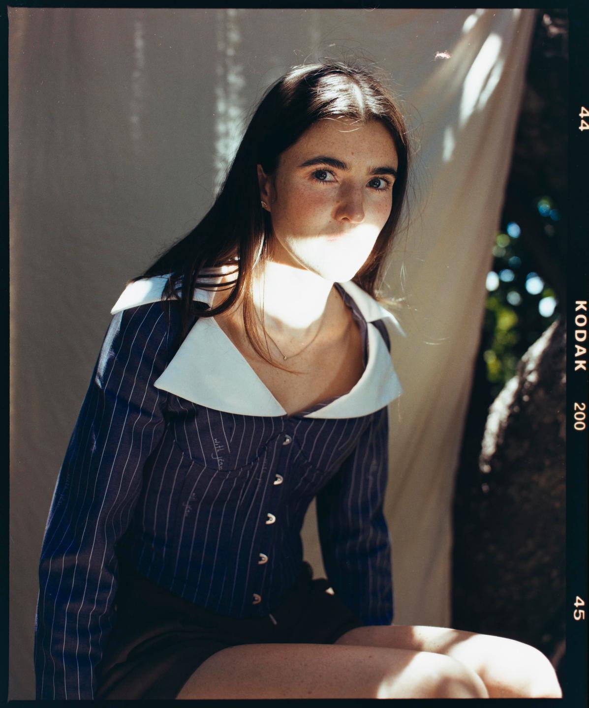 A woman with long hair and a striped shirt with a large white collar is sitting in dappled sunlight.