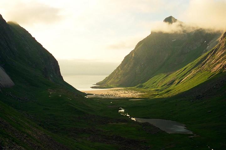 A valley with a stream and a lake leads to a sandy beach and the ocean, flanked by two large mountains under a cloudy sky.