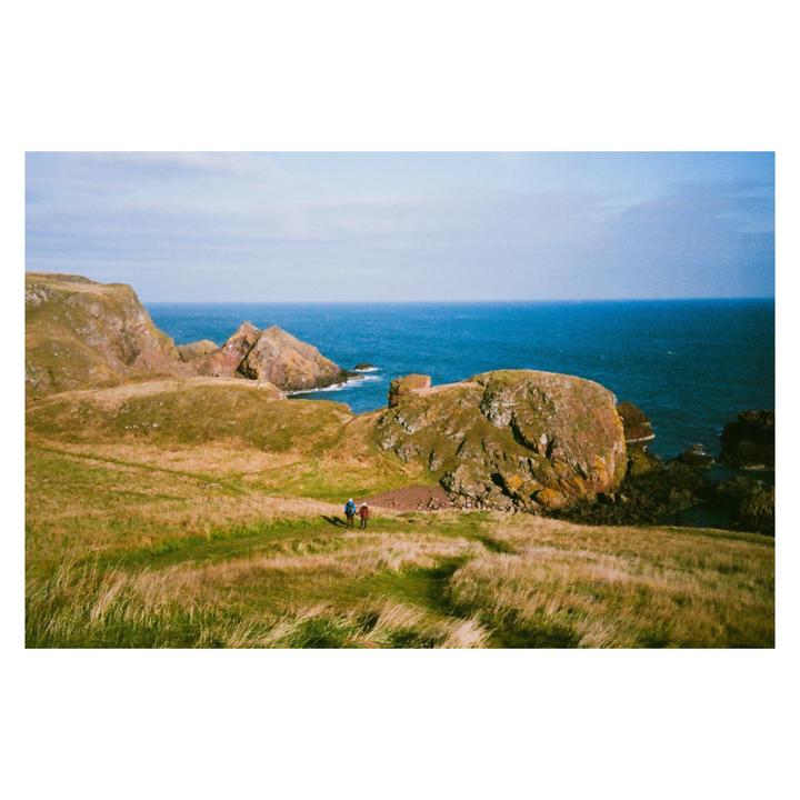 Two people walk along a grassy path on a cliff overlooking the ocean.