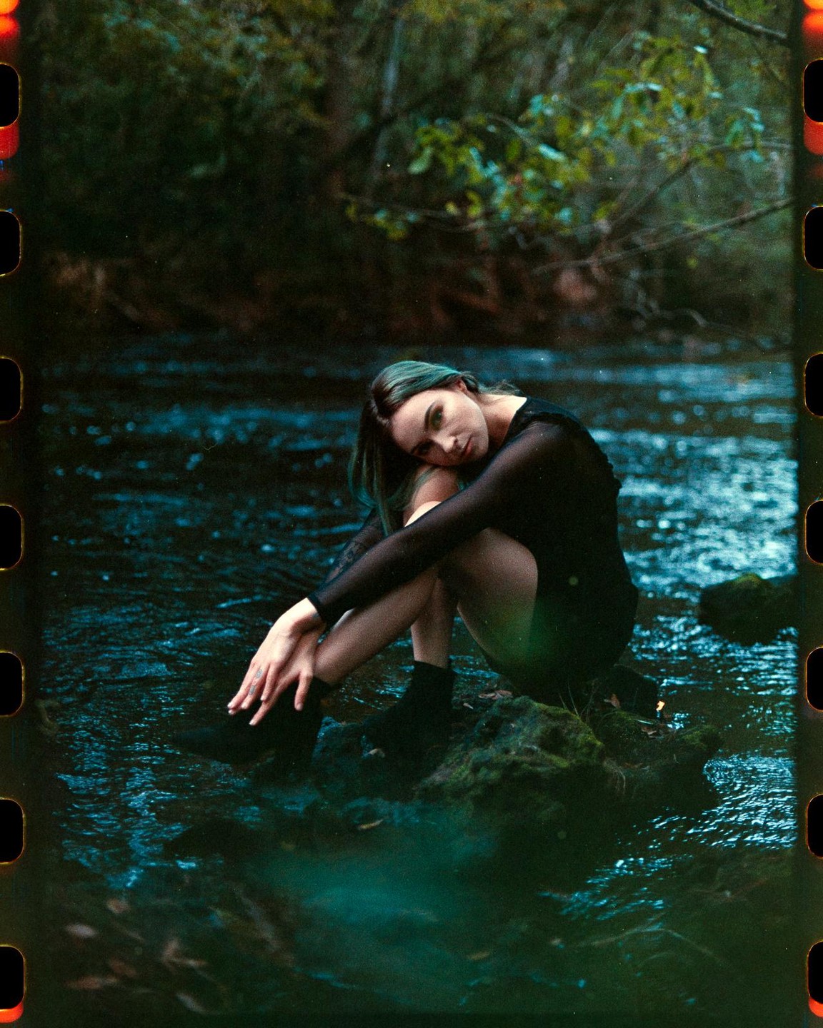 A woman with green hair sits on a mossy rock in a stream, looking towards the viewer.