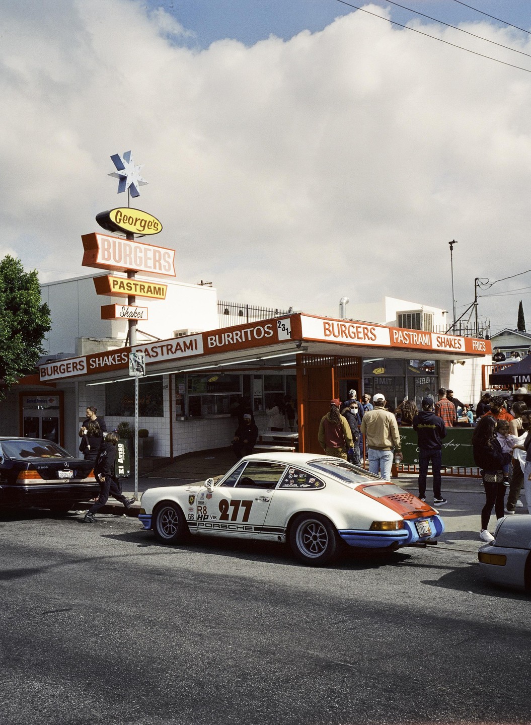 A white Porsche with racing decals is parked on a street in front of a burger restaurant with a large sign.