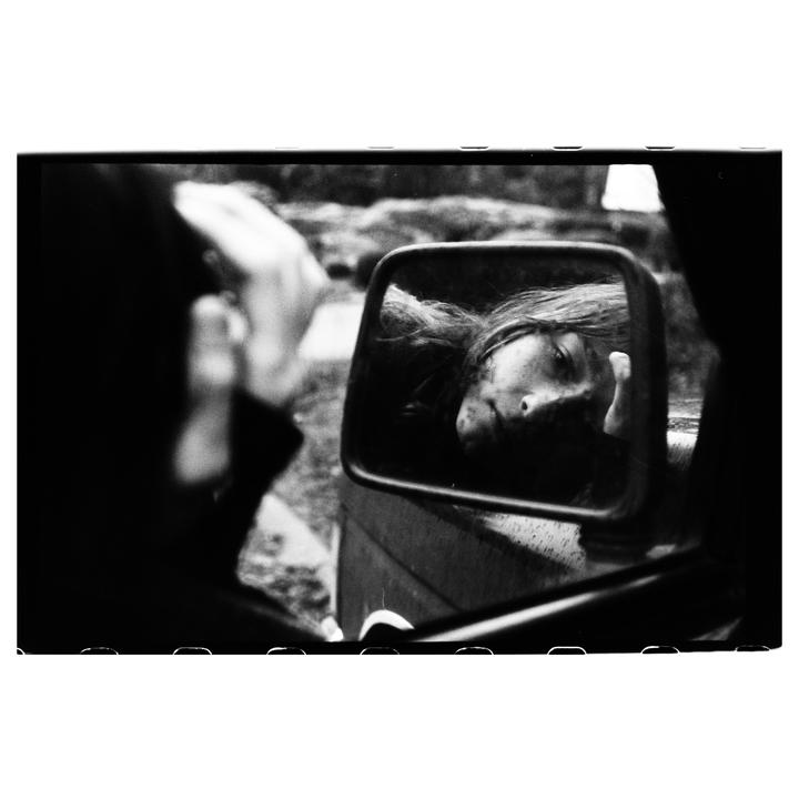 A woman's face is visible in a car's side mirror, with her hand resting on the mirror's frame.