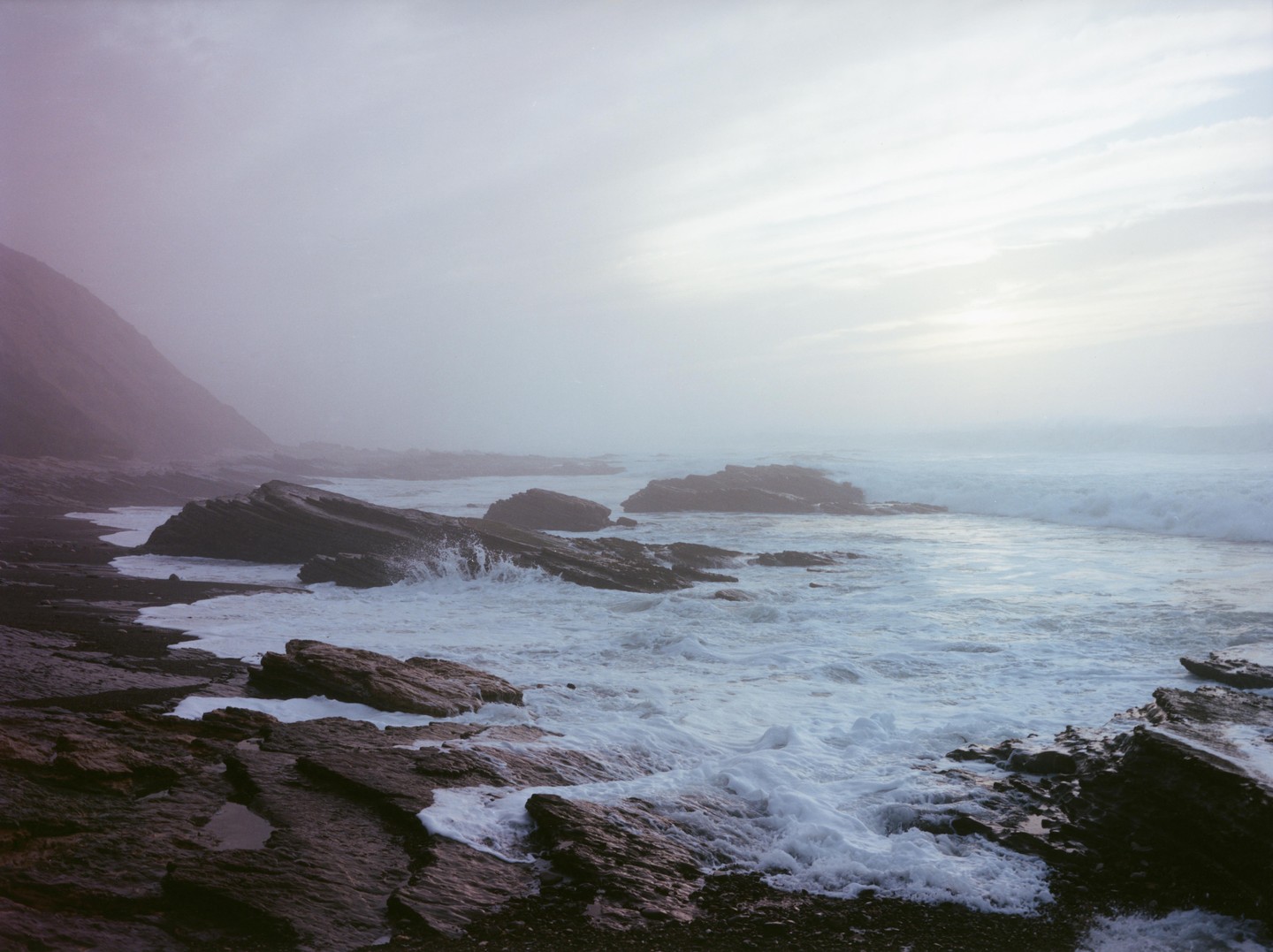 Waves crash against a rocky coastline under a hazy sky.
