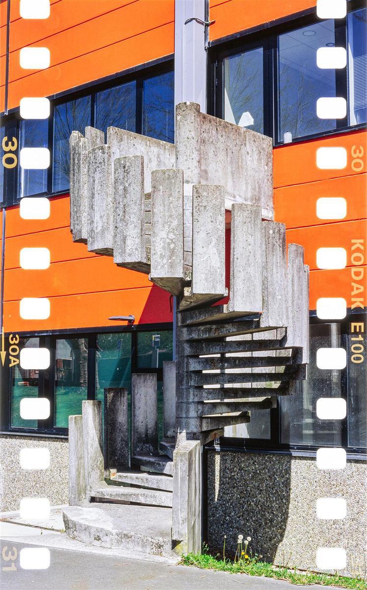 A concrete spiral staircase stands in front of an orange building with dark windows.