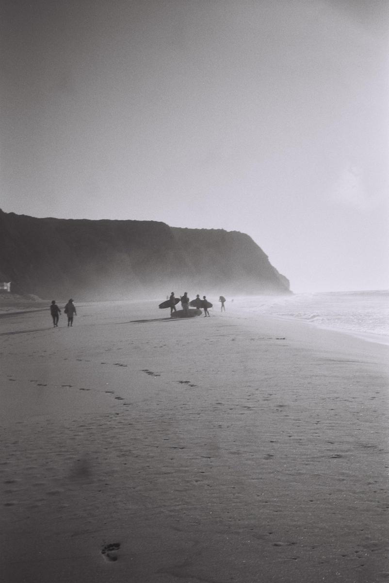 A group of surfers with their boards stands on a beach with a cliff in the background.
