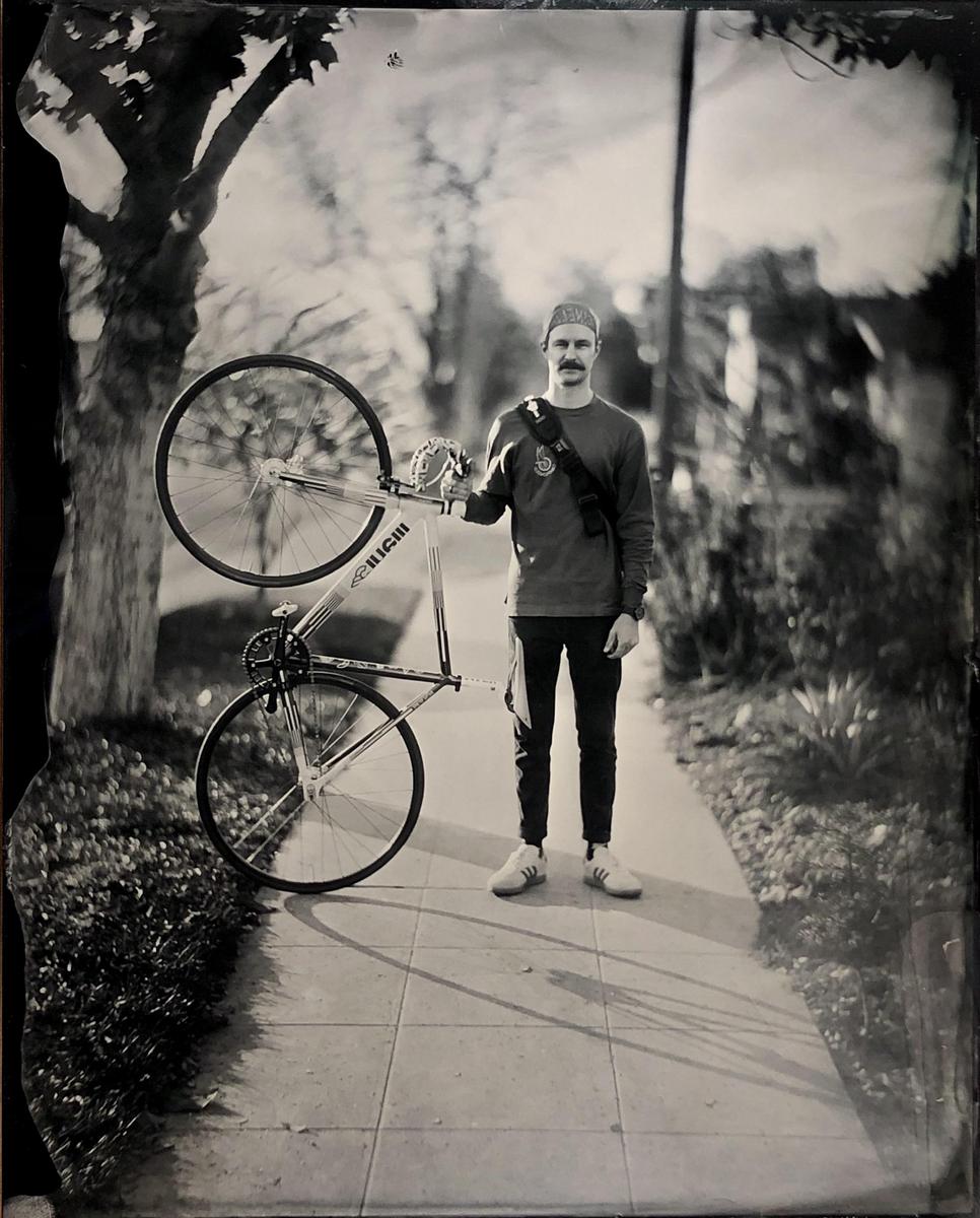 A man stands on a sidewalk holding a bicycle next to a tree.