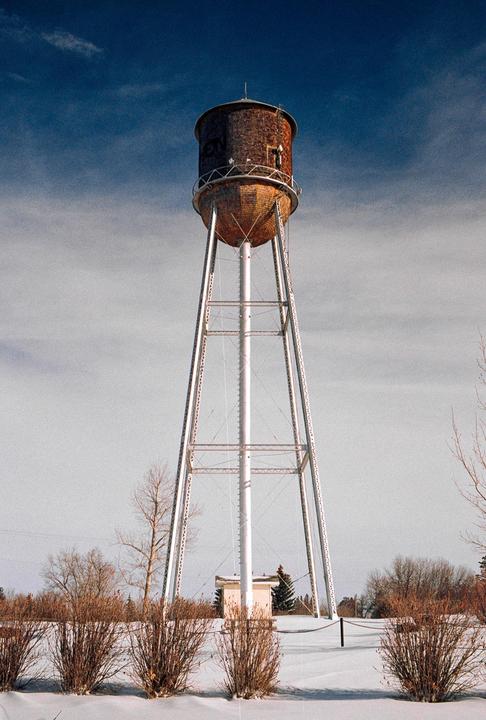 A tall water tower with a brick-like top stands over a snow-covered landscape with bare bushes and trees under a blue sky.