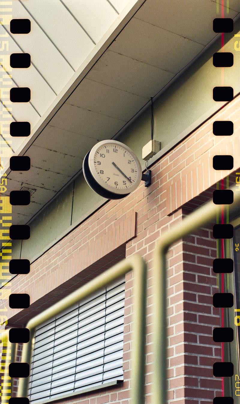 A clock is mounted on a brick wall above a window with blinds, with a yellow railing in the foreground.