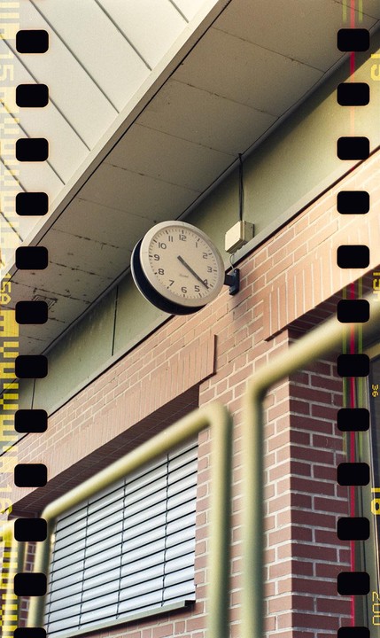 A clock is mounted on a brick wall above a window with blinds, with a yellow railing in the foreground.