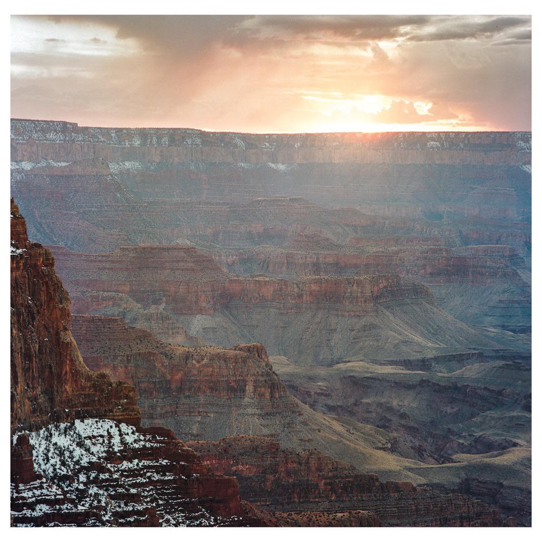 The sun sets over a vast canyon with snow-dusted ledges.