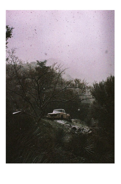 A vintage pickup truck is parked on a snowy hillside surrounded by trees under a hazy sky.