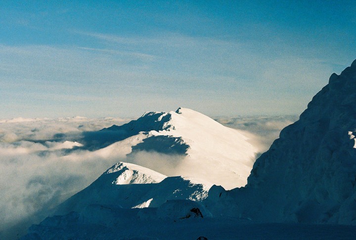 Snow-covered mountain peaks rise above a sea of clouds under a clear blue sky.