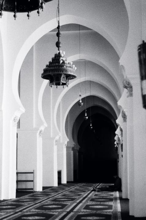 A person sits on a patterned carpet in a long hallway with arched columns and hanging lamps.