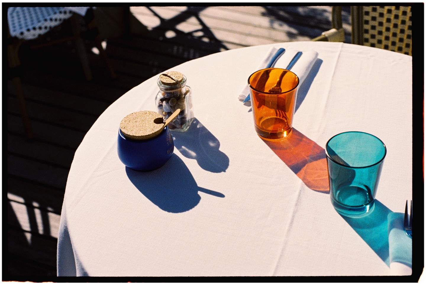 A white table is set with two colorful glasses, a jar of shells, and a blue sugar pot, all casting shadows.