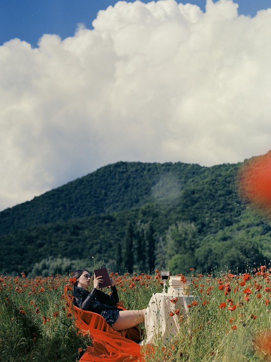 A woman in a black sequined top and red fabric is reading a book in a poppy field with a small table holding wine and books next to her, under a cloudy sky with a mountain in the background.