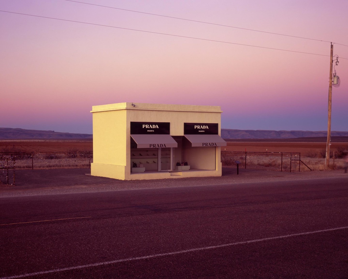 A small Prada store stands alone by a road under a colorful sky at sunrise.