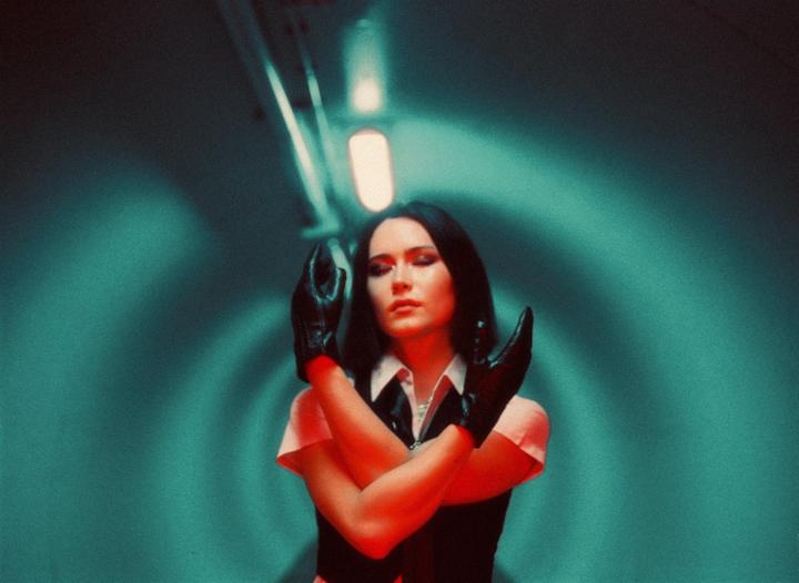 A woman with closed eyes and crossed arms wears black gloves and a black vest over a white collared shirt, illuminated by red light against a teal background.