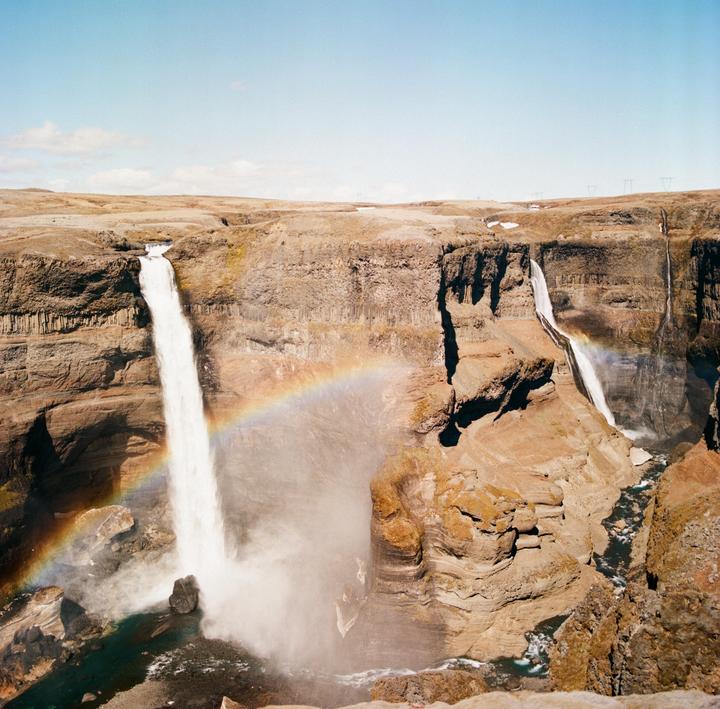 A rainbow arches over a large waterfall cascading into a canyon with a smaller waterfall visible in the distance.