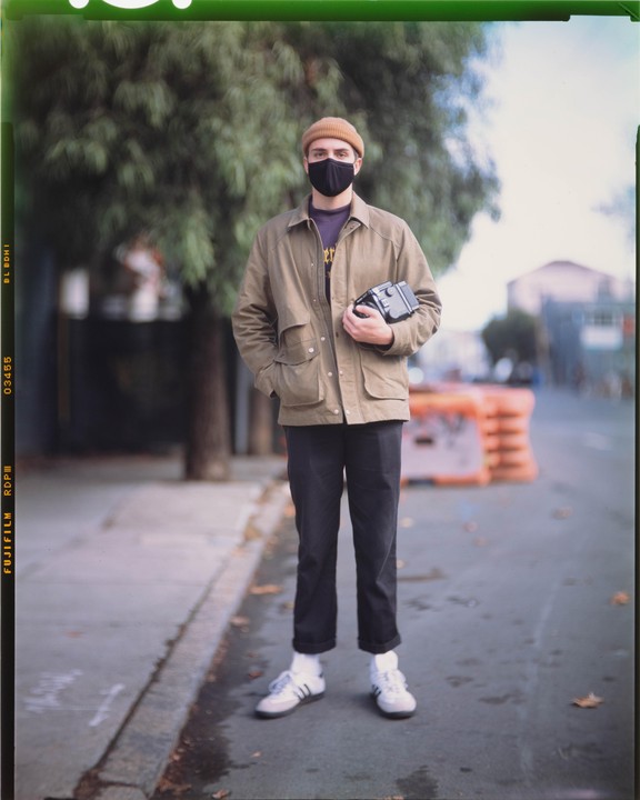 A man wearing a face mask and a beanie stands on a street holding a camera.