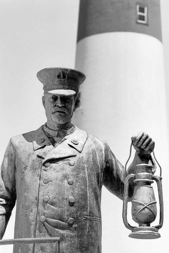 A statue of a lighthouse keeper holding a lantern stands in front of a lighthouse.