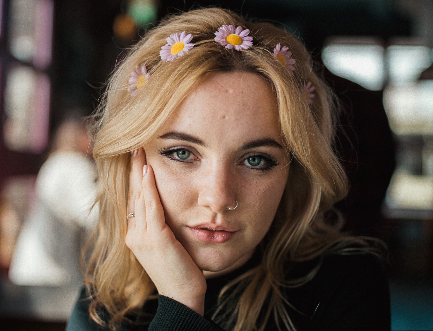 A woman with blonde hair and a flower headband looks directly at the viewer, resting her chin on her hand.