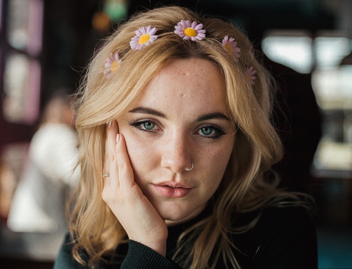 A woman with blonde hair and a flower headband looks directly at the viewer, resting her chin on her hand.