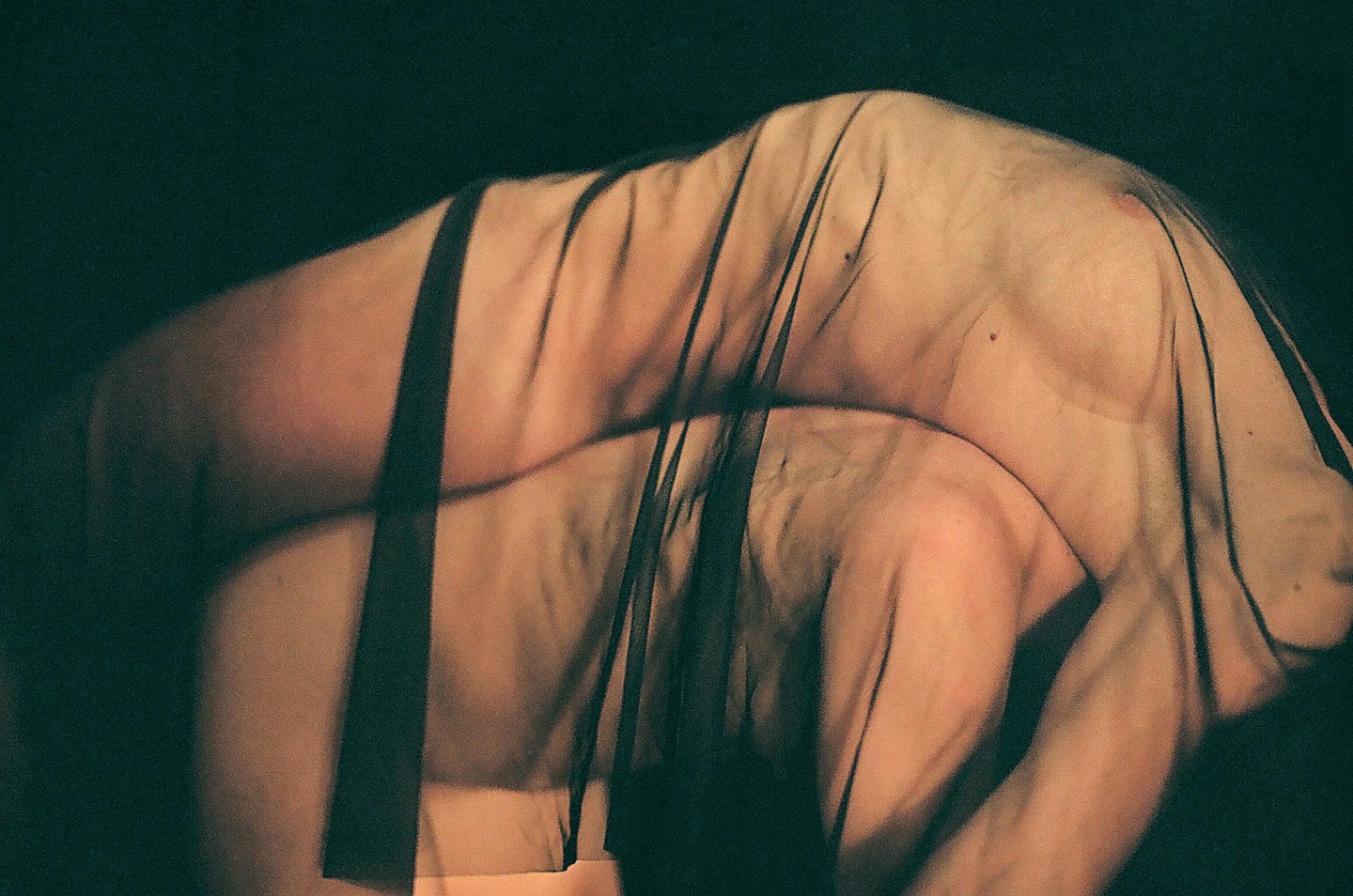 A person's body is draped in sheer black fabric against a dark background.