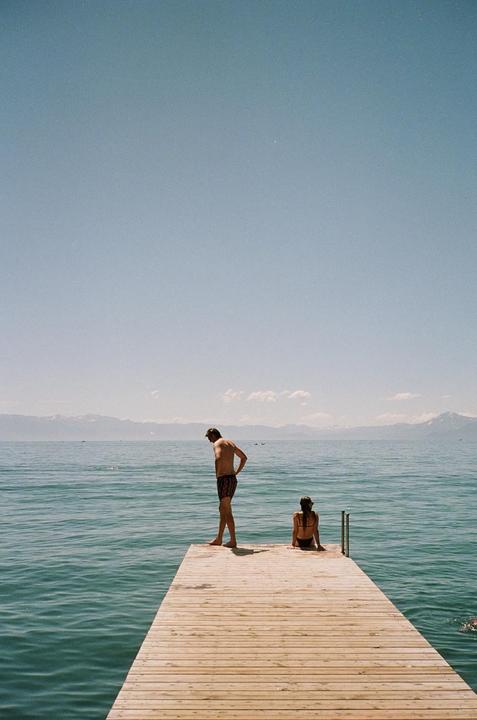 A man stands on a wooden pier looking down at the water while a woman sits at the end of the pier with mountains in the distance.