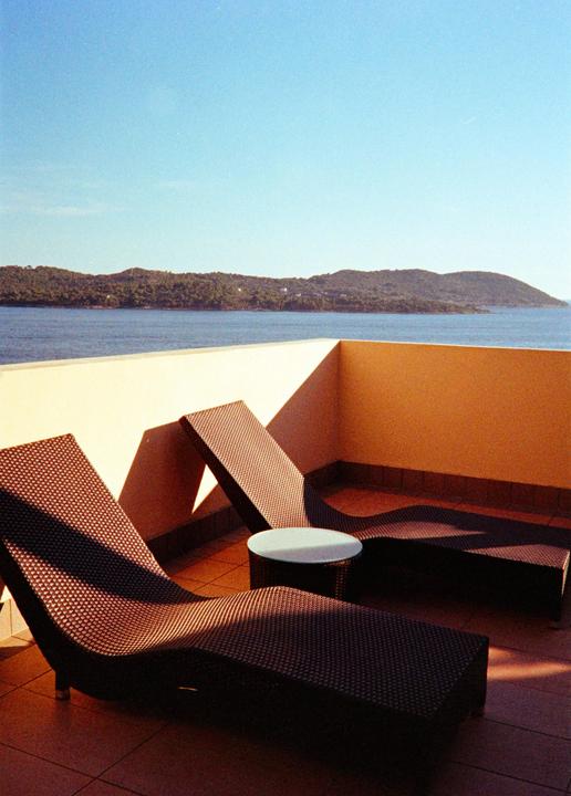 Two lounge chairs and a small table sit on a balcony overlooking a body of water and a distant forested island.